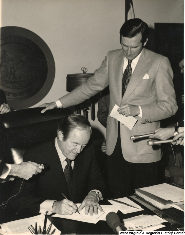 ["Secretary of State Jay Rockefeller watching Hubert Humphry sign a document while the press record what he is saying. Humphry was most likely in West Virginia when he was campaigning for President."]%